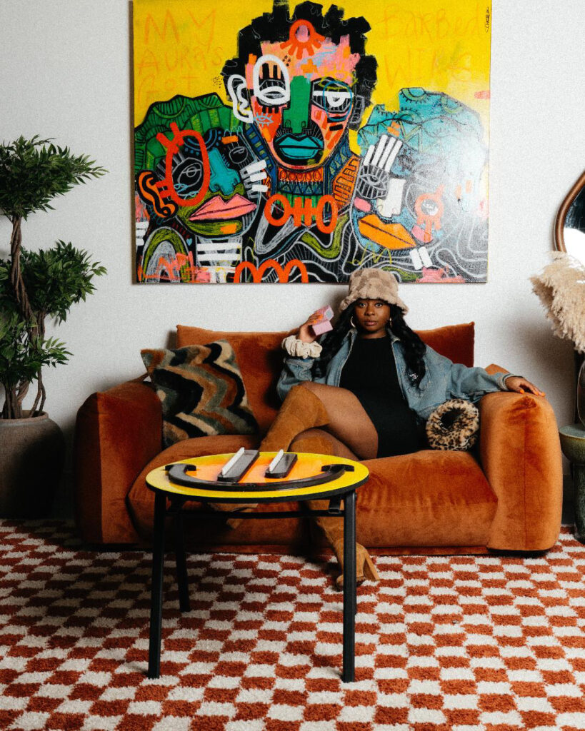 Artist Charmaine Chanakira on an orange velvet sofa beneath one of her paintings